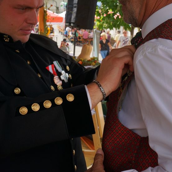 Ein uniformierter Mann mit Medaillen justiert die Krawatte eines Mannes in einem weißen Hemd. Der Hintergrund ist verschwommen mit Leuten und einem Zelt.