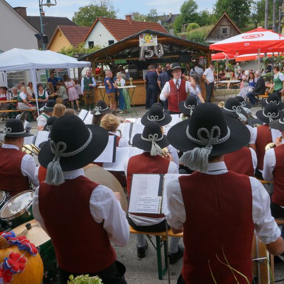 Eine Gruppe von Musikern in traditioneller Kleidung spielt Instrumente bei einem Open-Air-Festival, mit Zuschauern und einem Imbissstand im Hintergrund.
