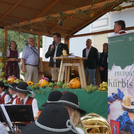 Eine Gruppe von Menschen ist in einem Zelt für eine Veranstaltung versammelt. Ein Mann spricht in ein Mikrofon an einem Podium. Musikinstrumente und Kürbisse sind auf dem Tisch vor ihnen.