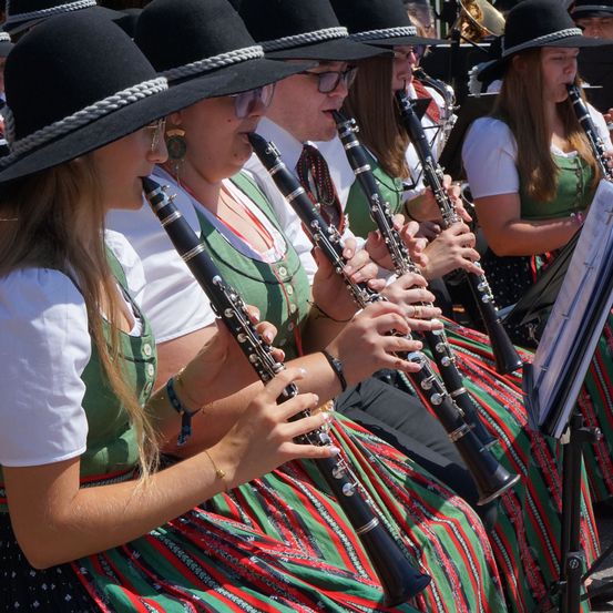Eine Gruppe von Frauen in traditioneller Kleidung spielt Klarinetten in einem Musikensemble. Sie sitzen in einer Reihe, wobei eine Frau im Vordergrund in ihre Klarinette bläst.