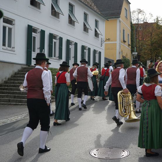 Eine Gruppe von Menschen in traditioneller Kleidung marschiert die Straße entlang und spielt Musikinstrumente. Die Gebäude im Hintergrund haben grüne Fensterläden.