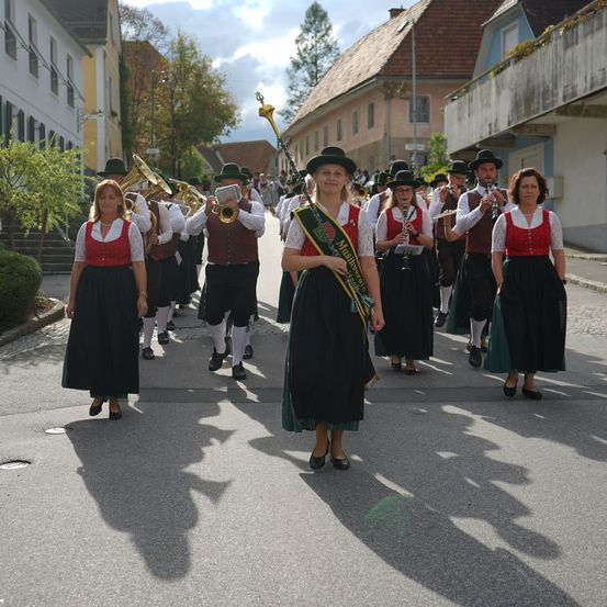 Eine Blaskapelle in traditioneller Kleidung tritt auf einer Straße auf, mit Gebäuden und Grünpflanzen im Hintergrund.