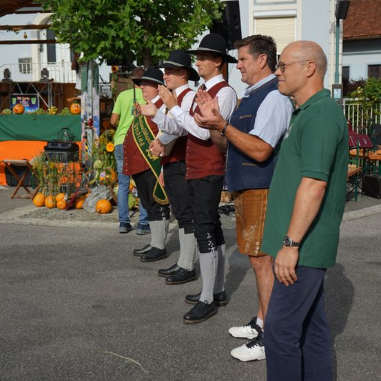 Fünf Männer in traditioneller deutscher Kleidung stehen in einer Reihe auf einer Straße und klatschen in die Hände. Dahinter ist eine Ausstellung von Kürbissen und Pflanzen aufgebaut.