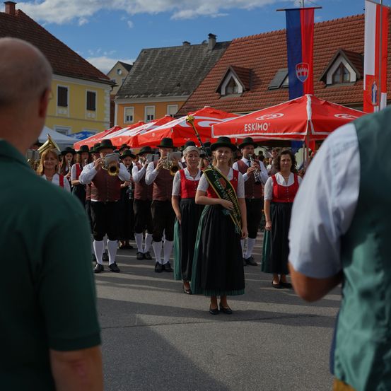 Ein Musikzug in traditioneller Kleidung tritt auf einem Stadtplatz unter roten Sonnenschirmen auf.
