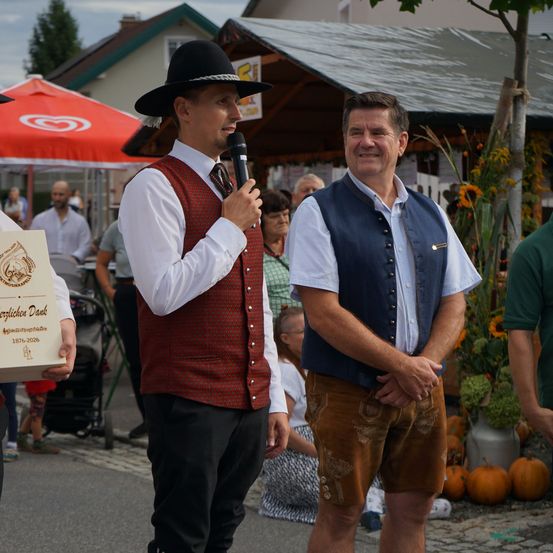 Zwei Männer in traditioneller bayerischer Kleidung stehen auf einer Straße, einer hält ein Mikrofon und der andere lächelt. Hinter ihnen ist ein Marktszene mit Kürbissen und einem roten Schirm zu sehen.