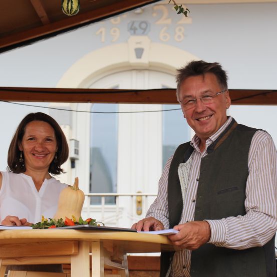 Ein Mann und eine Frau stehen hinter einem Tisch mit einem Kürbis und einigen Blumen darauf und lächeln in die Kamera.