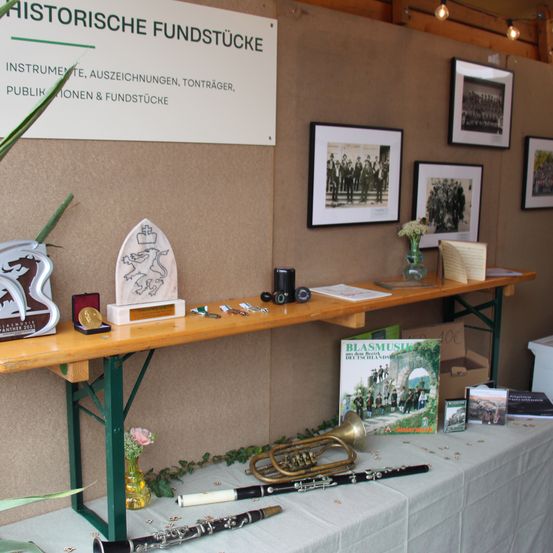 Eine Ausstellung historischer Blechblasinstrumente und Auszeichnungen auf einem Holzregal mit einem Schild, das 'Historische Fundstucke' besagt. Das Regal enthält eine Trompete, eine Klarinette, eine Medaille und eine Trophäe. Dahinter zeigt eine Wand gerahmte Fotografien und ein Schild.