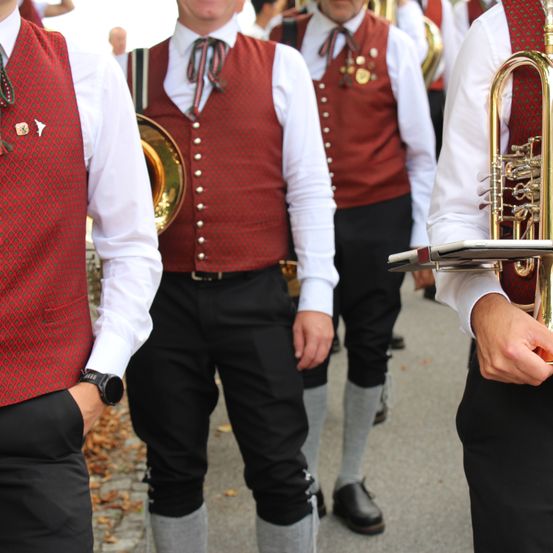 Eine Gruppe von Musikern in traditioneller Kleidung, die in einer Reihe stehen und Blechblasinstrumente halten. Der Mann auf der linken Seite trägt eine rote Weste mit einer schwarzen Uhr.