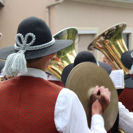 Ein Musiker in traditioneller Kleidung spielt ein Blechinstrument bei einer Parade, mit anderen Musikern und Instrumenten im Hintergrund.