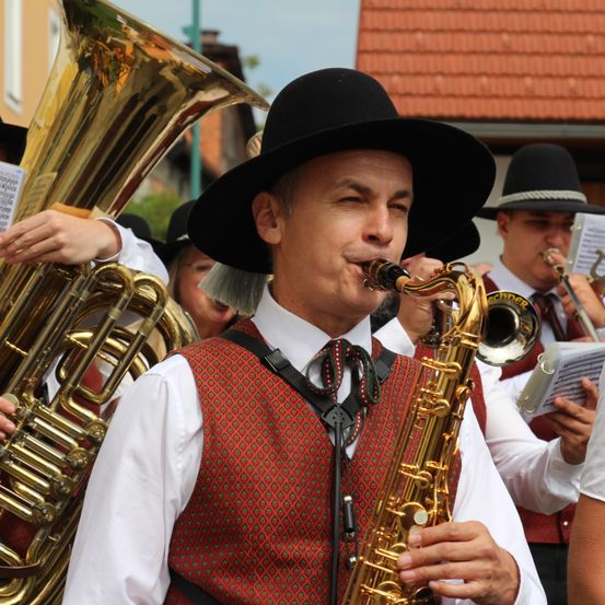 Ein Mann in einer roten Weste und schwarzem Hut spielt Saxophon bei einer Straßenaufführung mit anderen Musikern.