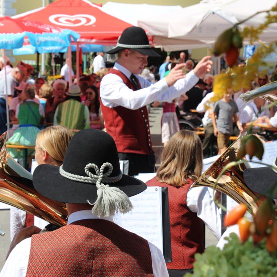 Ein Mann in traditioneller Kleidung dirigiert eine Brassband auf einem Festival, mit Zuschauern und Essensständen im Hintergrund.