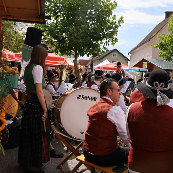 Eine Gruppe von Musikern in traditioneller Kleidung spielt im Freien, eine Frau hält einen Drumstick und ein Mann sitzt auf einer Bank vor einer Trommel.