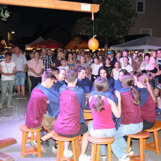 Eine Gruppe von Menschen in Lila sitzt um einen Tisch bei einer Veranstaltung im Freien, während eine Menge von hinten zuschaut. Die Umgebung ist nachts, mit Zelten und einer überhängenden Kürbis.