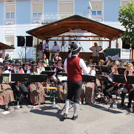 Ein Blasorchester spielt im Freien unter einem hölzernen Pavillon vor einem Gebäude mit Balkon. Die Musiker sind in traditioneller Kleidung gekleidet.