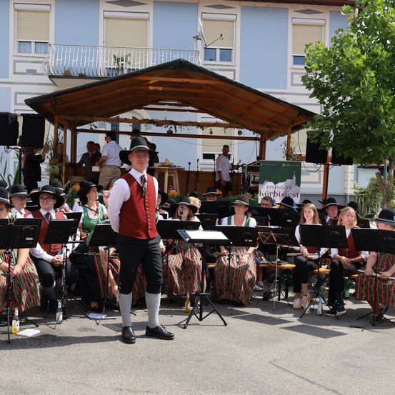 Eine Band in traditioneller Kleidung spielt auf der Bühne vor einem Gebäude mit Balkon und Lautsprechern.