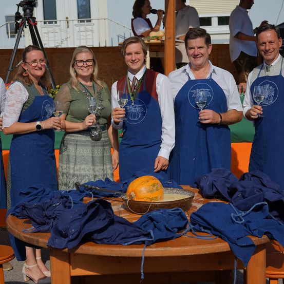 Fünf Personen in blauen Schürzen, lächelnd und mit Weinglas in der Hand, stehen um einen Holztisch mit einem Kürbis und blauem Tuch. Hinter ihnen sind Menschen in verschiedenen Aktivitäten beschäftigt.