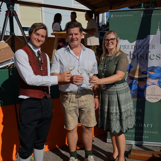 Drei Personen in traditioneller Kleidung halten Weinglas in der Hand und stehen vor einem Banner, das das Preding Kürbisfest bewirbt.