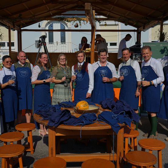 Eine Gruppe von Menschen in blauen Schürzen steht in einem Pavillon, lächelt und hält Weingläser. Ein Kürbis steht auf einem Tisch vor ihnen.