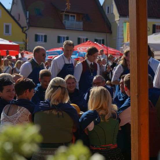 Eine Gruppe von Menschen in traditioneller Kleidung versammelt sich an einem Außenort, wahrscheinlich bei einem Festival. Einige tragen Schürzen und andere haben Schals. Im Hintergrund befinden sich Gebäude und rote Regenschirme.