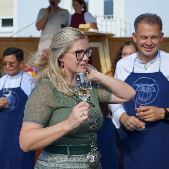Eine Frau in einem grünen Kleid hält ein Weinglas, während ein Mann in einer blauen Schürze neben ihr lächelt, mit anderen im Hintergrund.