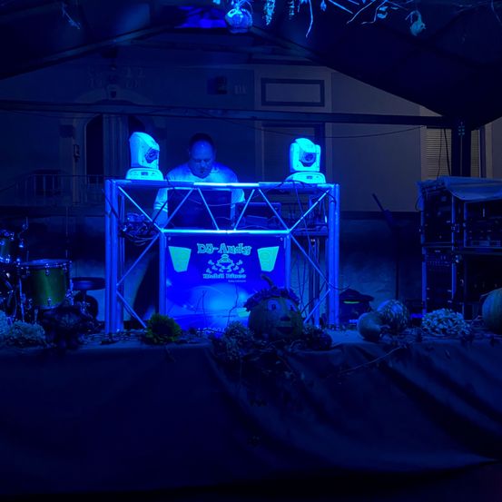 Ein Mann steht hinter einer DJ-Anlage auf einer Bühne, die von blauen Lichtern beleuchtet wird. Die Bühne ist mit Kürbissen und anderen Dekorationen geschmückt.