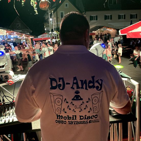 Ein Mann mit einem Shirt, auf dem DJ-Andy steht, steht an einer DJ-Konsole mit einer Menge im Hintergrund.