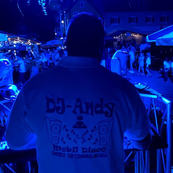 Ein Mann mit einem DJ-Andy-Shirt steht an einem DJ-Pult bei einer blau beleuchteten Outdoor-Veranstaltung, mit einer Menge im Hintergrund.