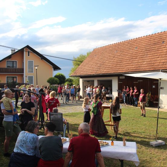Eine Gruppe von Menschen versammelt sich in einem Hof mit Häusern im Hintergrund, einige stehen, andere sitzen an einem Tisch mit Essen.