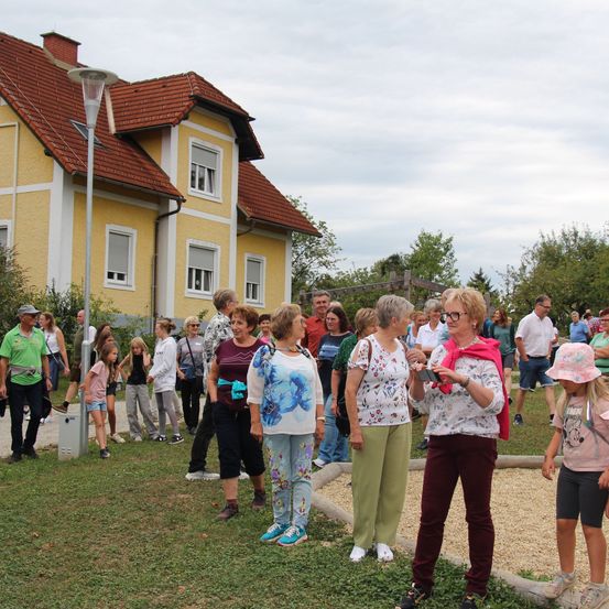 Eine Gruppe von Menschen, einschließlich Erwachsener und Kindern, steht auf einer Rasenfläche vor einem gelben Haus mit rotem Dach. Einige tragen Brillen und haben Taschen bei sich.