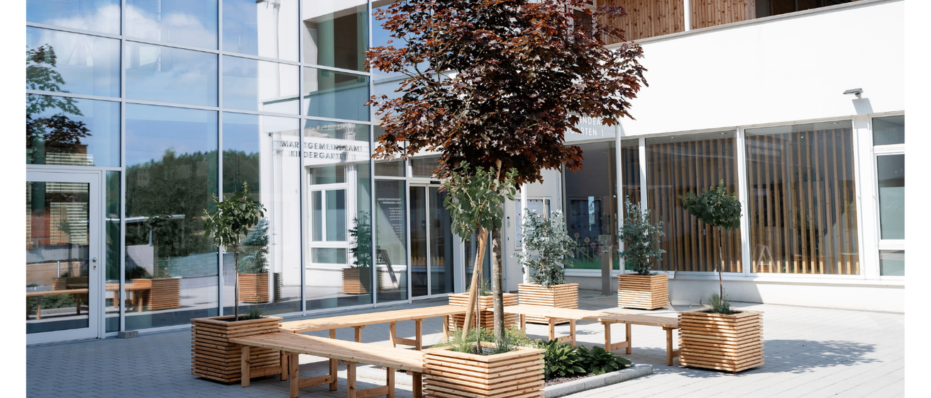 Ein modernes Gebäude mit Glaswänden und Holzakzenten, das einen Innenhof mit Holzbänken, einem zentralen Baum und mehreren Topfpflanzen aufweist.