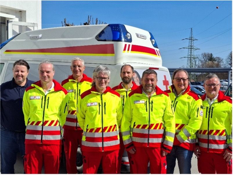 A group of people in yellow and red uniforms stand in front of an ambulance. The uniforms feature reflective stripes and red crosses.