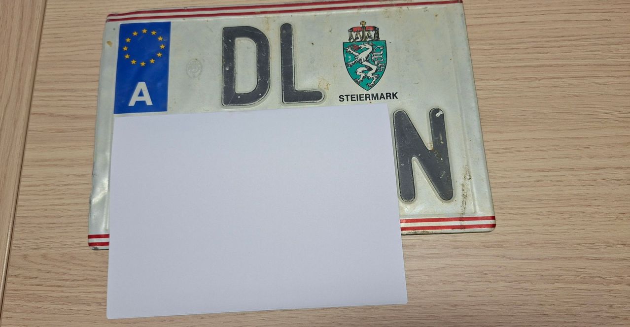 Ein österreichisches Autokennzeichen mit den Buchstaben DL und dem Wappen von Steiermark, teilweise von einem weißen Blatt Papier bedeckt.