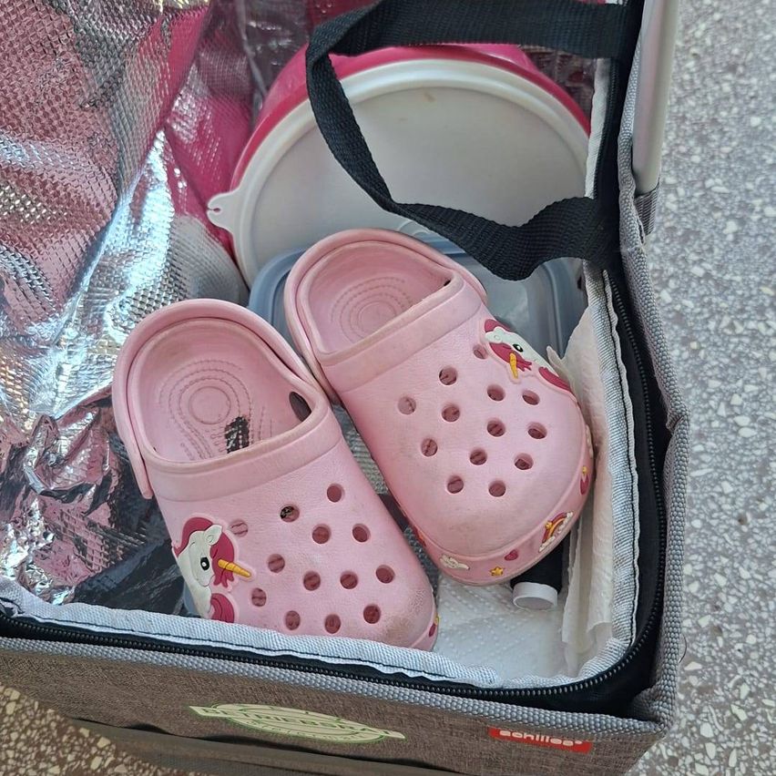 A pair of pink unicorn-themed Crocs inside a transparent plastic bag, next to a pink plastic container and a black strap.