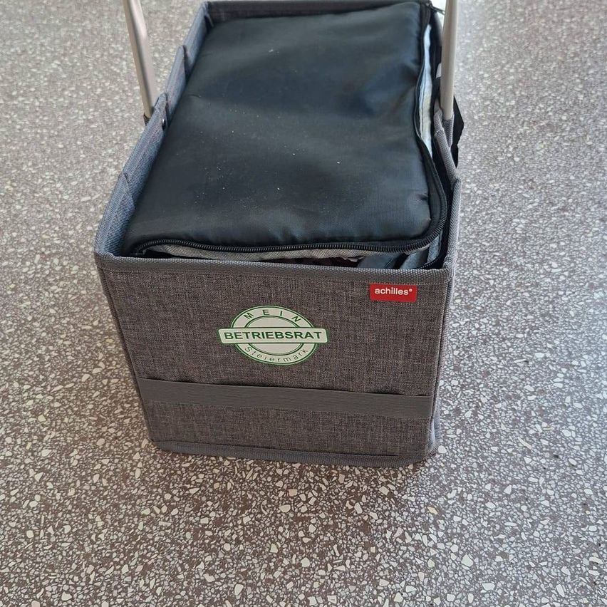 A gray shopping basket with a handle and a black cushion inside, featuring a red label with the word 'achilles' and a green logo reading 'BETRIEBSRAT'. The basket is placed on a speckled floor.