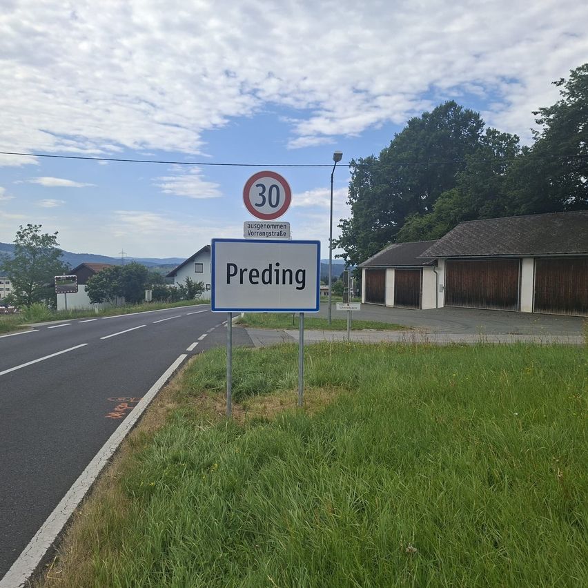 A street sign for Preding with a 30 km/h speed limit is on the roadside. Behind the sign, there are houses and a building with a garage. The surroundings are filled with trees and grass.