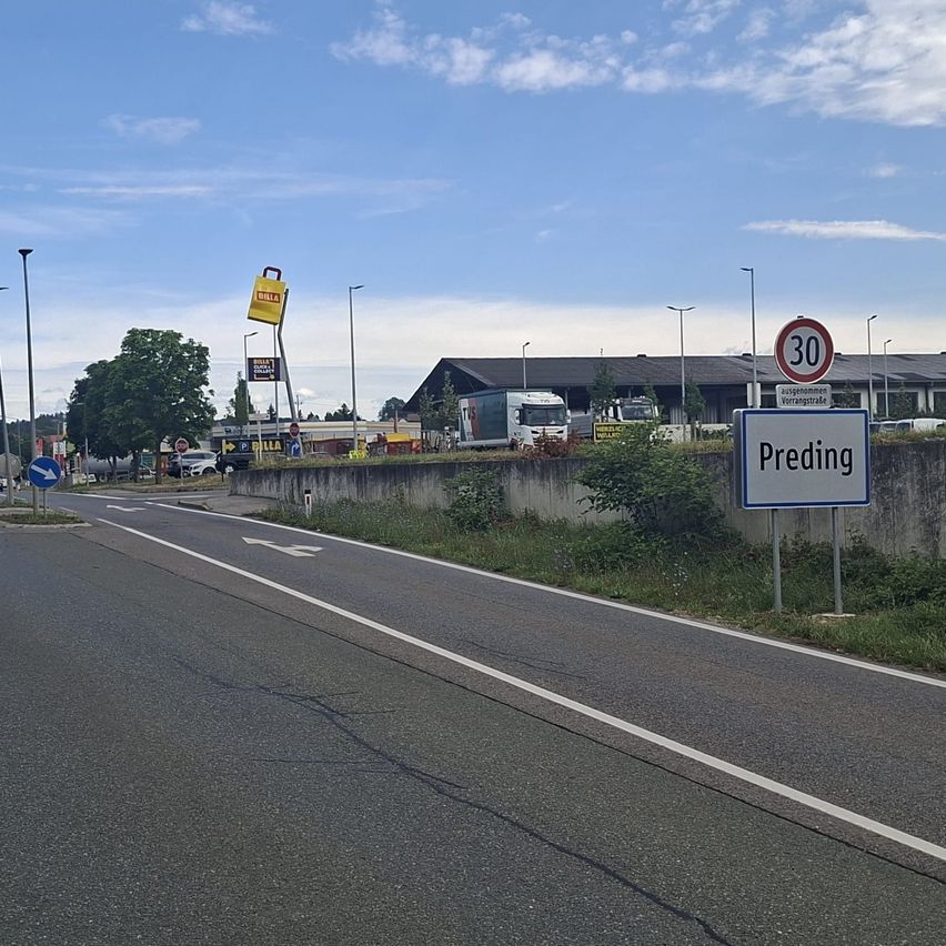 An empty road with a speed limit sign of 30 km/h. There is a sign indicating Preding. Several buildings are visible in the background.