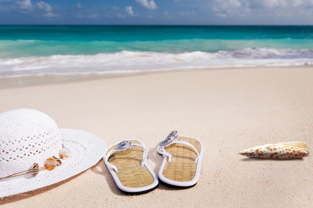 A pair of flip-flops adorned with jewels lie on a sandy beach, near a white hat and seashells, with the ocean waves in the background.
