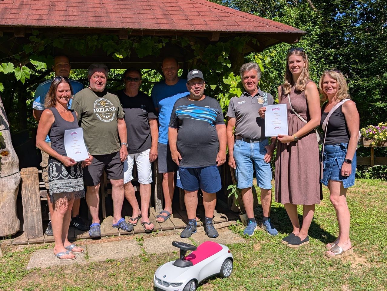A group of adults stand under a gazebo, smiling and posing for a photo. They hold certificates and some wear sunglasses. A toy car is on the ground.