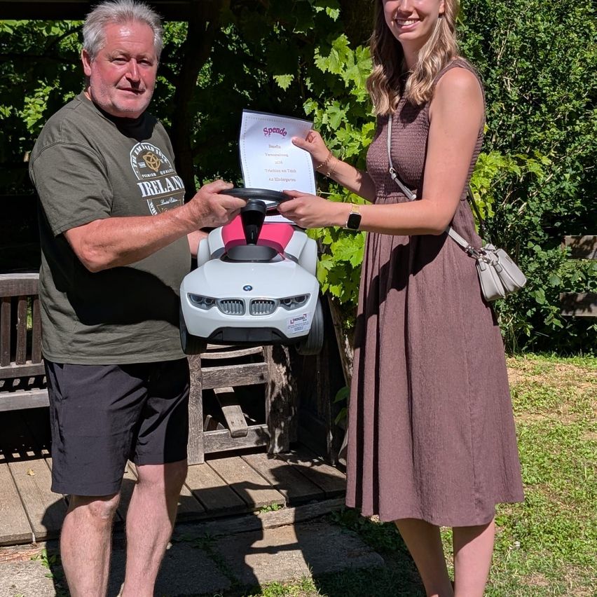 A man and woman are standing in a garden, holding a toy car and a certificate. The man smiles, wearing a green shirt, and the woman smiles brightly. They are surrounded by greenery.