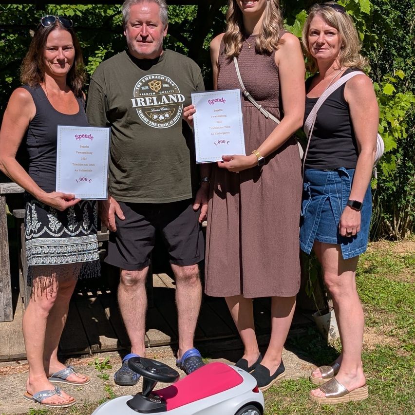 Four people are standing in an outdoor area, holding certificates. A woman holds a certificate with 1,000 euros. Another woman holds a certificate with 'Spende'. A man is wearing a shirt that says 'Premium Quality Ireland'. A woman is wearing a watch and sandals.