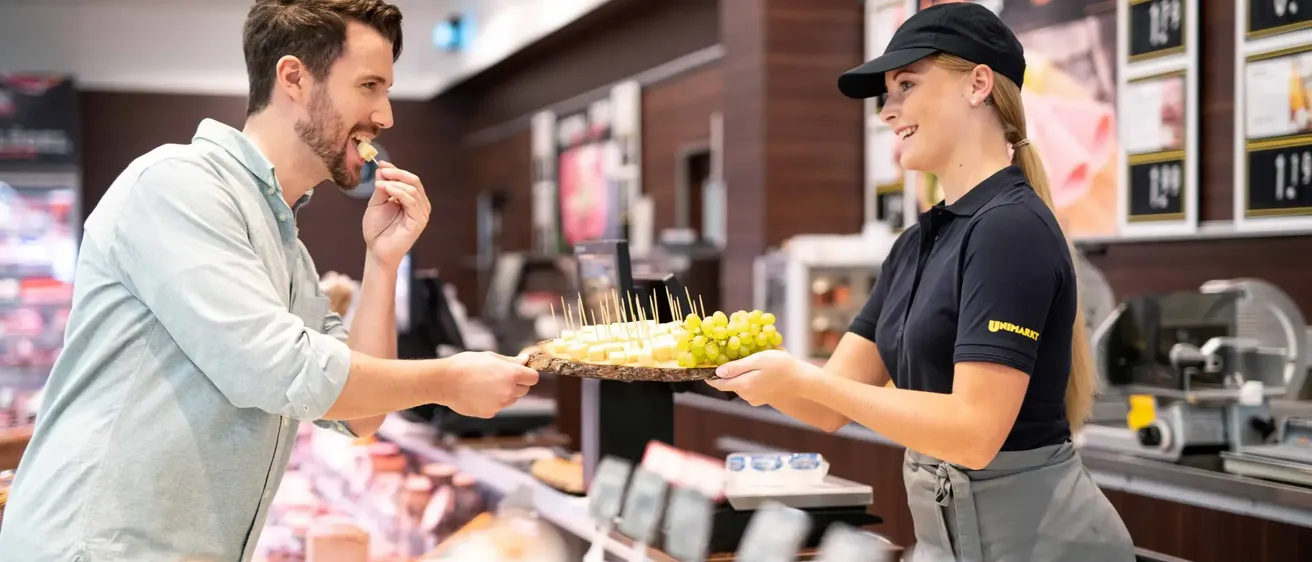 Ein Mann lächelt, während er einen Snack an einer Ladentheke isst. Eine Frau in einer schwarzen Kappe und einem schwarzen Hemd reicht ihm einen Teller mit Essen.