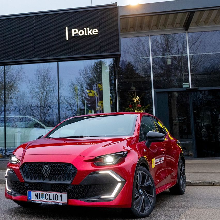 Ein roter Renault Clio steht vor einem Geschäft mit dem Schild 'Polke'. Das Auto hat das Kennzeichen 'MI CLIO 1'. Bäume und ein weiteres Auto sind in den Glasfenstern reflektiert.