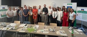 Eine Gruppe von Menschen steht in einem Konferenzraum, einige halten Urkunden. Sie sind von Tischen mit verschiedenen Gegenständen umgeben. Der Raum hat ein modernes Design mit weißen Wänden und einem grünen Banner auf der rechten Seite.