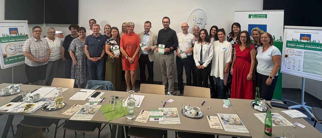 Eine Gruppe von Menschen steht in einem Konferenzraum, einige halten Urkunden. Sie sind von Tischen mit verschiedenen Gegenständen umgeben. Der Raum hat ein modernes Design mit weißen Wänden und einem grünen Banner auf der rechten Seite.