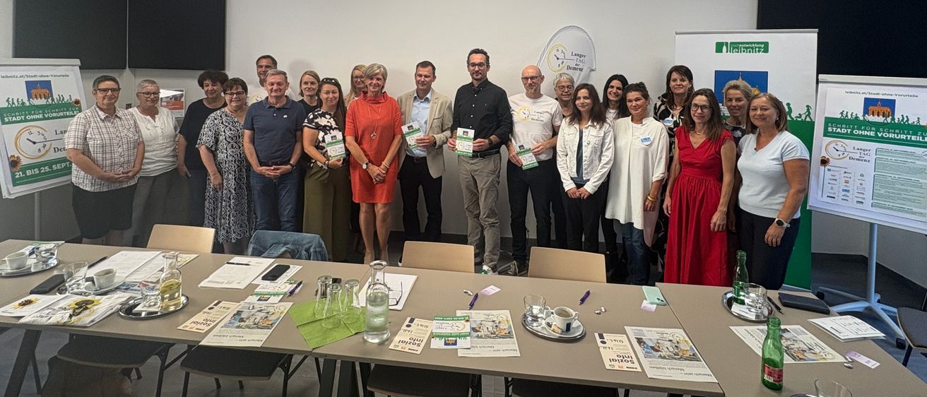 Eine Gruppe von Menschen steht in einem Konferenzraum, einige halten Urkunden. Sie sind von Tischen mit verschiedenen Gegenständen umgeben. Der Raum hat ein modernes Design mit weißen Wänden und einem grünen Banner auf der rechten Seite.