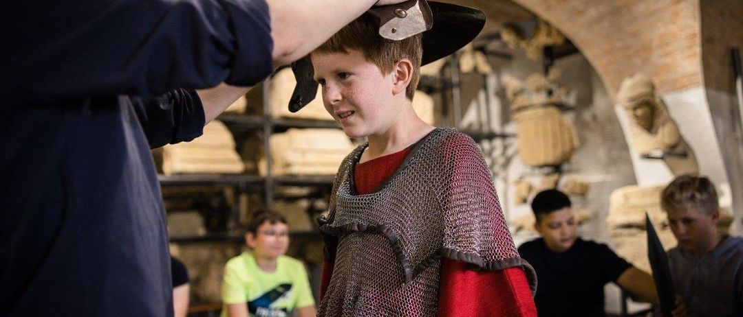 Ein junger Junge wird von einem Erwachsenen mit einem Metallhelm ausgestattet, während einige Leute im Hintergrund sitzen und zusehen.