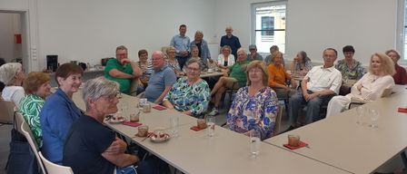 Eine Gruppe älterer Menschen sitzt um einen Tisch herum, wahrscheinlich in einer Besprechung. Einige tragen Brillen, andere Mützen. Sie scheinen miteinander zu sprechen. Der Raum ist weiß, und es gibt ein Fenster mit Jalousien.