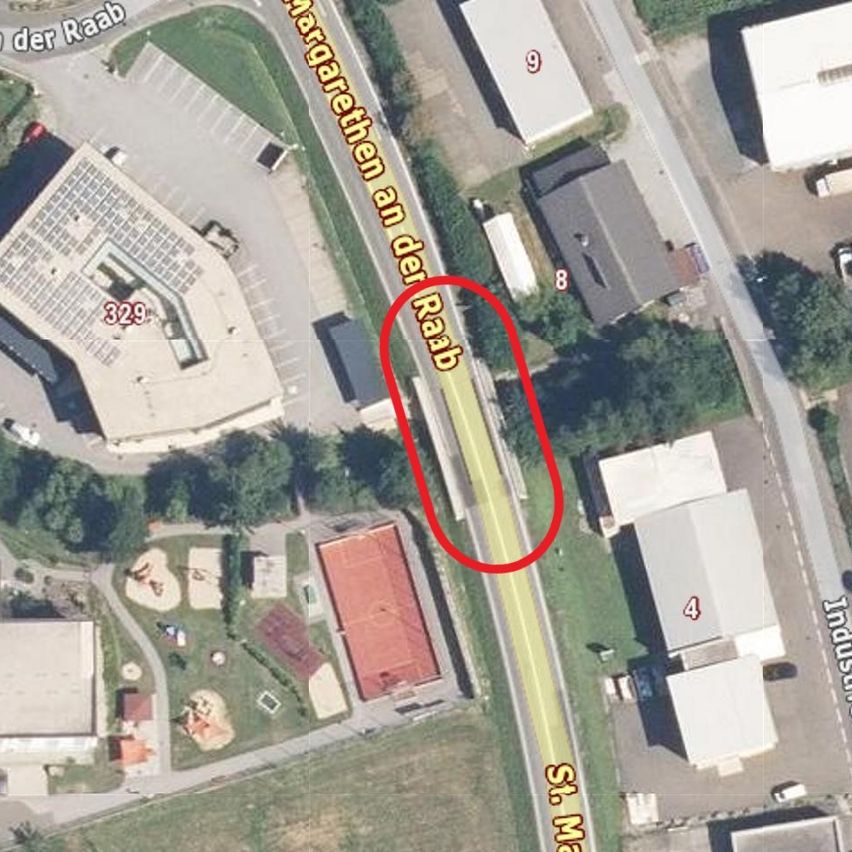 Luftaufnahme einer Straße mit mehreren Gebäuden. Ein roter Kreis hebt einen bestimmten Abschnitt hervor. Ein Gebäude hat Solarpanels, und es gibt verschiedene andere Strukturen wie Tennisplätze und einen Spielplatz.