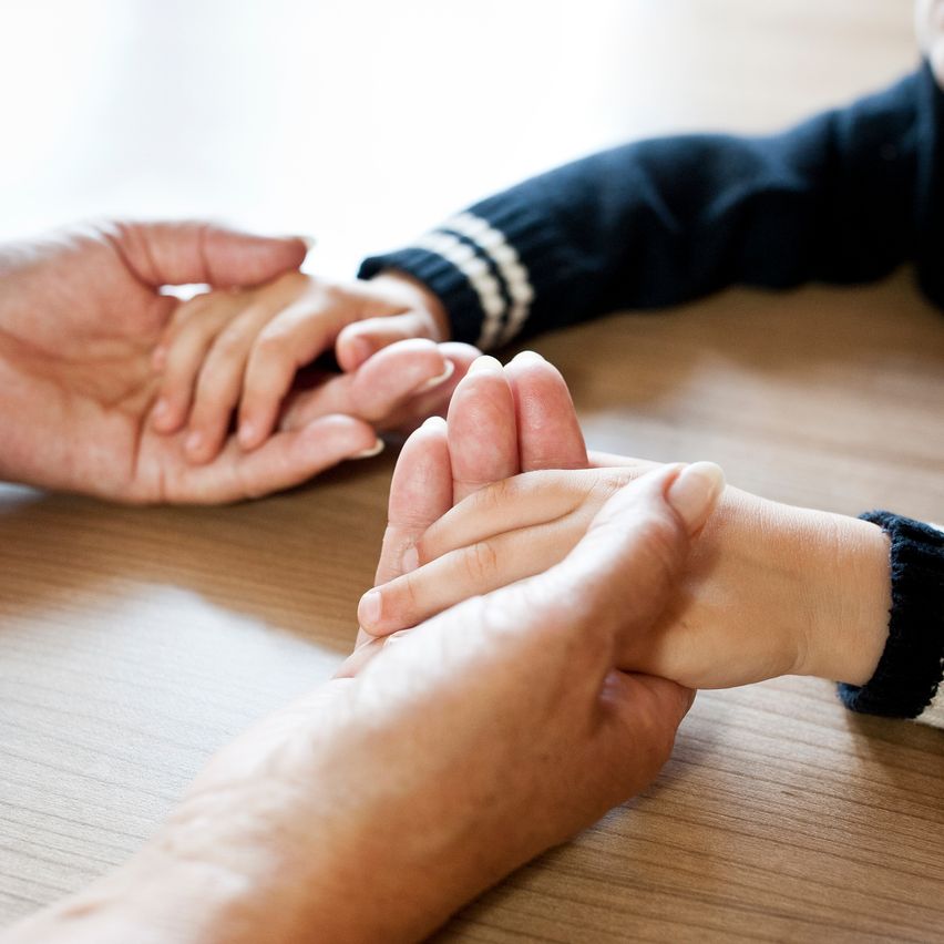 Ein Nahaufnahme von zwei Händen, die sich halten, eine erwachsene und eine von einem Kind, mit ineinander verschlungenen Fingern auf einem Holztisch.
