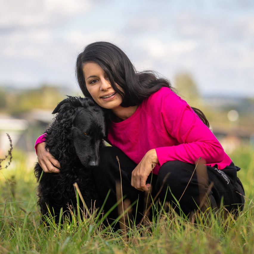 Eine Frau mit langen schwarzen Haaren, die ein rosa langes Oberteil und schwarze Hose trägt, sitzt in einem Grasfeld mit einem schwarzen Hund auf ihrem Schoß.